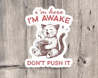 May include: A sticker featuring a yawning cat holding a coffee mug. The text reads "I'm here, I'm awake, don't push it" in red. The cat is brown and white, with steam rising from the mug.