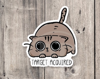 Puede incluir: Una ilustración de dibujos animados de un gato marrón con ojos grandes y una expresión de sorpresa. El gato está mirando al espectador y tiene el texto "TARGET ACQUIRED" debajo.