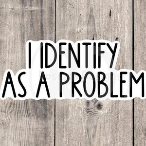 May include: White sticker with black text that reads "I identify as a problem". The sticker is on a wood background.