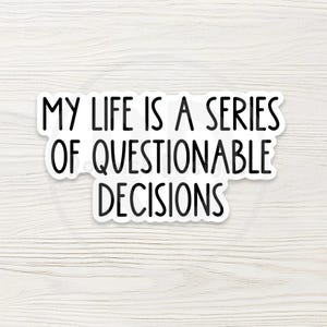 May include: A white sticker with black text that reads "MY LIFE IS A SERIES OF QUESTIONABLE DECISIONS." The sticker has a black border and is set against a light wood grain background.