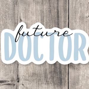 May include: A white sticker with the words "future DOCTOR" in a combination of script and block letters. The word "future" is in black script, and "DOCTOR" is in light blue block letters. The sticker is on a wooden background.