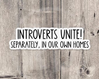 May include: A white sticker with the text "INTROVERTS UNITE! SEPARATELY, IN OUR OWN HOMES" in black, on a wooden background. The sticker has a simple, bold font, and the background is a light brown with wood grain.