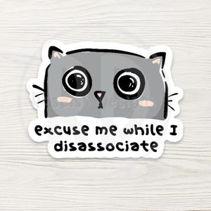 May include: A gray cat sticker with large, expressive eyes and pink cheeks. The sticker has the text "excuse me while I disassociate" in a playful font. The design is outlined in black on a white background.