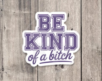 May include: A white sticker with the text "BE KIND of a bitch" in purple block letters and script. The sticker is on a wooden background.