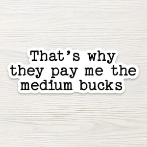 May include: A white sticker with a black border and the text "That's why they pay me the medium bucks" in a bold, sans-serif font. The sticker is on a light wood-grain background.