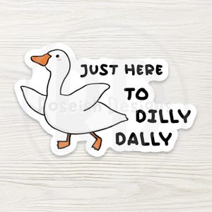May include: A white sticker with a cartoon goose, orange beak and feet, and the text "JUST HERE TO DILLY DALLY" in black. The sticker is on a white and light brown wooden surface.