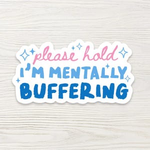 May include: A white sticker with the text "please hold I'm mentally buffering" in pink, blue, and dark blue. The sticker has a white border and is decorated with blue starbursts. The sticker is on a white wooden surface.