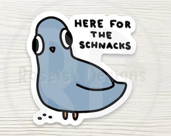 May include: A sticker featuring a cartoon pigeon in shades of blue, with large eyes and a speech bubble that reads "HERE FOR THE SCHNACKS". The pigeon is outlined in black and has small orange feet. A few black specks are near the pigeon.