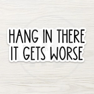 May include: A white sticker with a black border and the text "HANG IN THERE IT GETS WORSE" in a bold, sans-serif font. The sticker is set against a light wood grain background.
