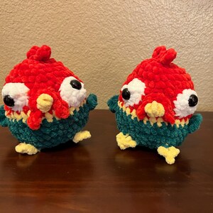May include: Two handmade crochet creatures with red, white, yellow, and teal details. The plush toys have large, black eyes and a whimsical design. The creatures are sitting on a dark brown surface.
