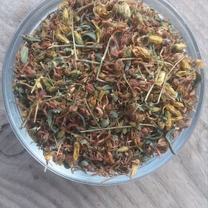 May include: A glass bowl filled with dried herbs and flowers, including yellow, brown, and green pieces. The bowl is sitting on a wooden surface.