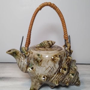May include: A ceramic teapot shaped like a seashell with a brown woven handle. The teapot has a light brown glaze with green accents and a seashell on the lid.