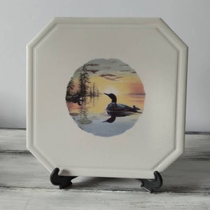 May include: A white ceramic tile with a raised octagonal border. The tile features a circular image of a loon swimming in a lake at sunset. The image is surrounded by a white border.