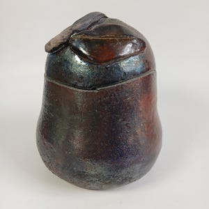 May include: A dark brown ceramic jar with a lid. The lid has a textured, abstract design. The jar has a mottled, iridescent glaze.
