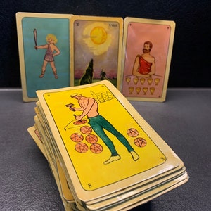 Puede incluir: Una baraja de cartas de tarot con ilustraciones coloridas. Las cartas est&aacute;n apiladas una encima de la otra y muestran una variedad de im&aacute;genes, incluyendo un hombre con un martillo, un lobo y un hombre con una copa. Las cartas son amarillas con n&uacute;meros negros en las esquinas.