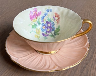 May include: A vintage teacup and saucer set with a floral design. The teacup is white with a gold rim and a floral pattern in shades of pink, blue, yellow, and green. The saucer is a pale pink color with a gold rim and a scalloped edge.