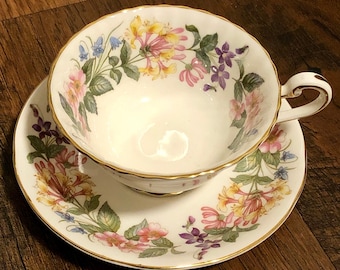 May include: A white teacup and saucer with a floral pattern. The pattern features pink, yellow, blue, and purple flowers with green leaves. The cup and saucer have a gold trim.