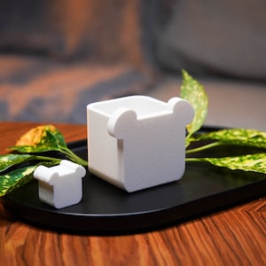 Peut inclure: Deux pots en céramique blanche en forme d'ours. Le plus grand pot a une base carrée et le plus petit pot est un cube. Les deux pots sont sur un plateau noir sur une table en bois.