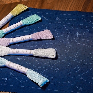 May include: Five spools of embroidery floss in various pastel colours, including light blue, green, yellow, pink, and lavender. The floss is arranged in a rainbow pattern on a dark blue fabric with a white star pattern.