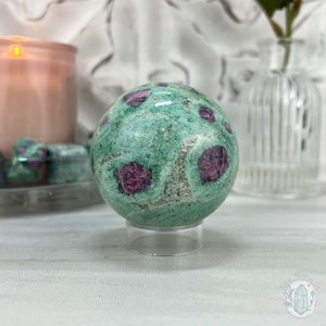 May include: Green and pink gemstone sphere on a clear stand. The sphere has a speckled appearance with pink inclusions.