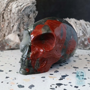 May include: A carved skull sculpture with a polished finish. The skull is primarily dark gray with red and gray marbling. The sculpture has a smooth, glossy surface and is displayed on a speckled surface.