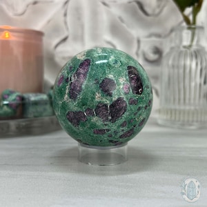 May include: A green and pink sphere with a speckled pattern, sitting on a clear stand. The sphere is made of a natural stone, possibly a type of jasper or aventurine.
