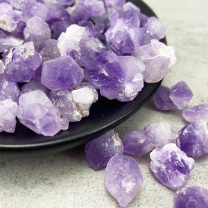 May include: A collection of small, purple amethyst crystals in various shapes and sizes. The crystals are arranged on a black plate and some are scattered on a gray surface.