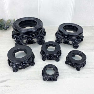 May include: A collection of six black decorative stands. Each stand features a circular top with a detailed, ornate base. The stands vary in size, with the largest at the top and the smallest at the bottom. The stands are made of a glossy black material.