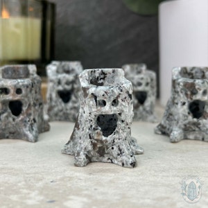 May include: Four small, white and gray stone carvings of tree stumps with carved faces. The carvings have a black hole for a mouth and two small holes for eyes.