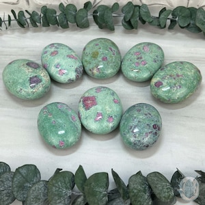 May include: Eight smooth, oval-shaped green and pink stones with red flecks. The stones are arranged in a circle on a white surface.