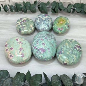 May include: Six smooth, oval-shaped green and pink stones with a speckled pattern. The stones are arranged in a row on a light gray surface.