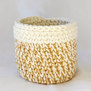 Peut inclure: Un panier en crochet avec un dessus blanc et un fond doré. Le panier est fait d'un fil épais et texturé et a une forme ronde.