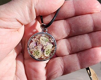 Puede incluir: Un collar con un colgante redondo de plata que contiene flores blancas y rosas prensadas en resina. El collar tiene un cordón negro.