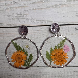 May include: A pair of handmade earrings featuring pressed orange flowers, green leaves, and small white and pink flowers encased in clear resin. The earrings have a silver-colored, irregular circular frame and a round stud.