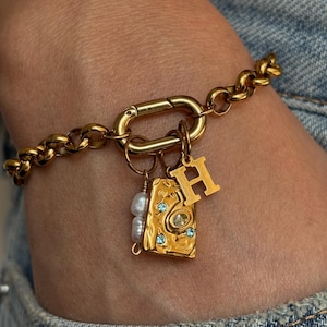 May include: A gold-toned chain link bracelet featuring a large oval clasp. Attached to the clasp are three charms: a letter H, a rectangular gold-toned pendant with an irregular surface, small blue crystals, and a small cluster of white pearls.
