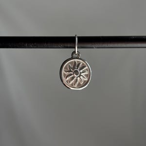 May include: A silver charm with a sun design. The charm is round and has a raised sun with rays emanating from it.