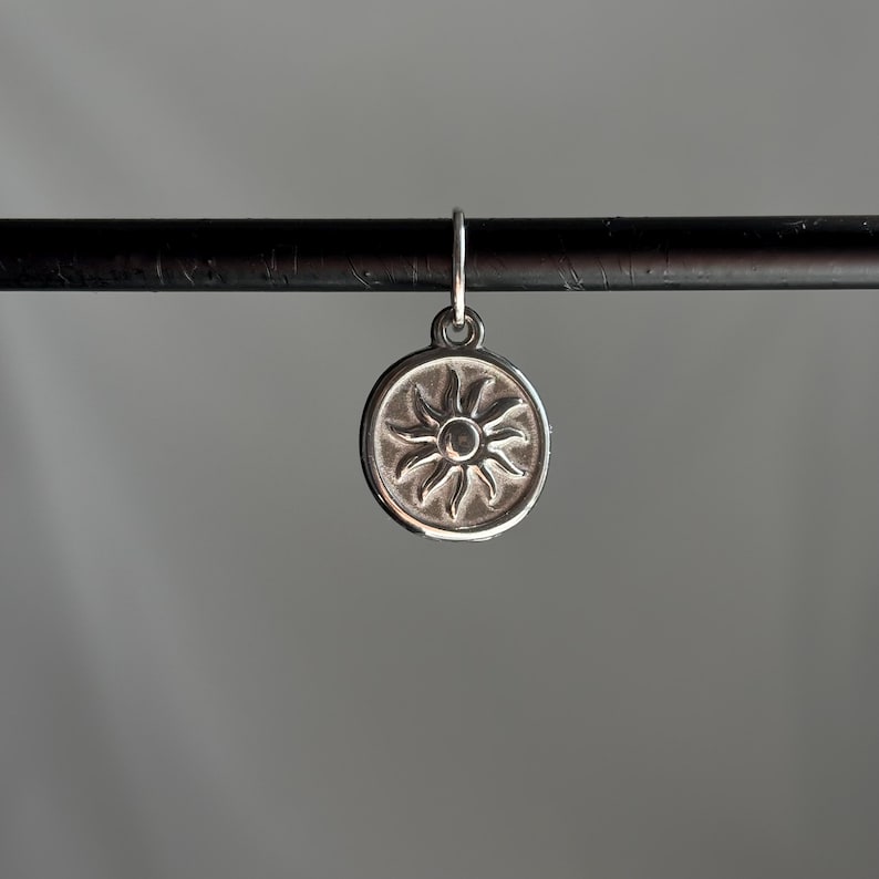 May include: A silver charm with a sun design. The charm is round and has a raised sun with rays emanating from it.