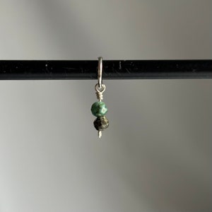 May include: A silver charm with two green gemstone beads. The top bead is a light green and the bottom bead is a darker green with black flecks.