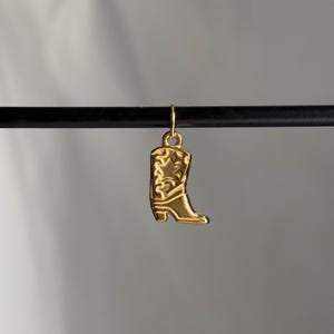 May include: A gold-colored cowboy boot charm with a star design. The charm is hanging from a black rod.