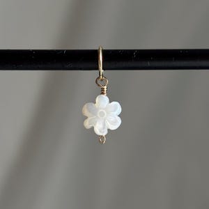 Puede incluir: Un delicado charm de oro con una flor blanca con un diseño tallado. El charm está unido a un bucle de oro.