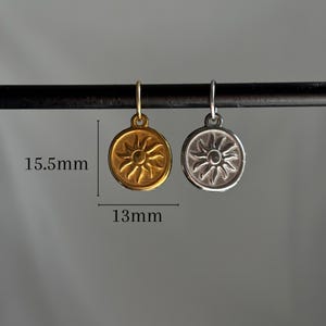 May include: Two sun charm pendants, one gold and one silver, hanging from a black rod. The charms are 13mm in diameter and 15.5mm tall.