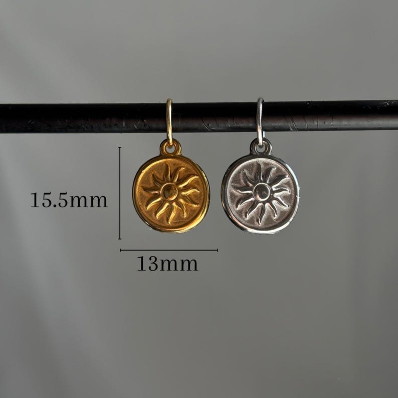 May include: Two sun charm pendants, one gold and one silver, hanging from a black rod. The charms are 13mm in diameter and 15.5mm tall.
