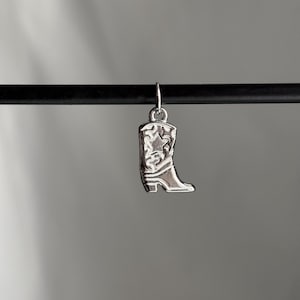 Pode incluir: Charme de bota de cowboy de prata com design de estrela. O charme está pendurado em um cordão preto.