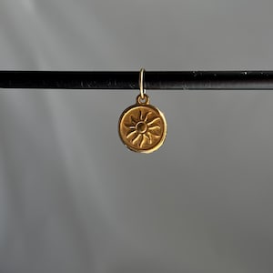 Op de afbeelding: Gouden bedel met een zonontwerp. De bedel is rond en heeft een reliëfzonontwerp met stralen die uit het midden komen.