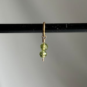 Może przedstawiać: Złoty charm z dwoma zielonymi fasetowanymi koralikami zwisającymi z niego.