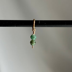 Puede incluir: Un amuleto de oro con dos cuentas de piedras preciosas verdes. El amuleto está unido a un anillo de oro.