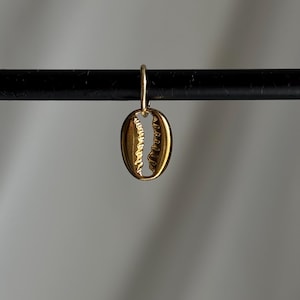 May include: A gold-coloured seashell charm pendant. The charm is shaped like a cowrie shell with a small hole at the top for attaching to a necklace or bracelet.