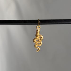 Puede incluir: Un colgante de serpiente dorada con detalles intrincados cuelga de un pequeño lazo. La serpiente está enrollada, con una superficie texturizada. El colgante está suspendido sobre un fondo gris neutro.