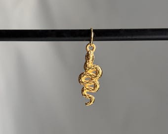 May include: A gold-colored snake pendant with intricate detailing hangs from a small loop. The snake is coiled, with a textured surface. The pendant is suspended against a neutral gray background.