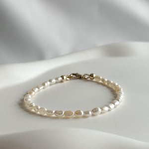 May include: Delicate pearl bracelet featuring small, irregularly shaped white pearls strung together. The bracelet has a gold-colored clasp and connecting elements. The pearls are a creamy white color, and the bracelet is displayed on a white surface.